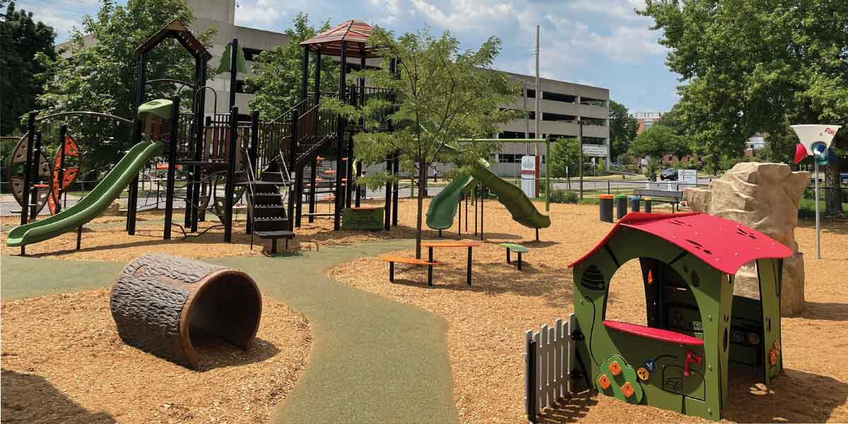 Playground containing green pathways leading to a large fort-style playground structure, and playhouse and log tunnel in the foreground