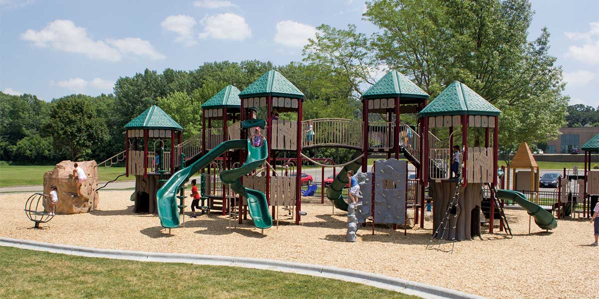 Large Minnesota outdoor playground with green, Burgundy, and tan. Using a playfort and nature theme, there are board slat style safety enclosures on the towers, and molded equipment in the shape of rocks and trees.